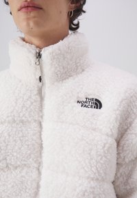 Valkoinen fleece-takki korkealla kauluksella, vetoketjukiinnitys ja logo. Pehmeä tekstuuri, jonka pinnassa on paksu tyynykuviointi ja mustia yksityiskohtia rinnassa.