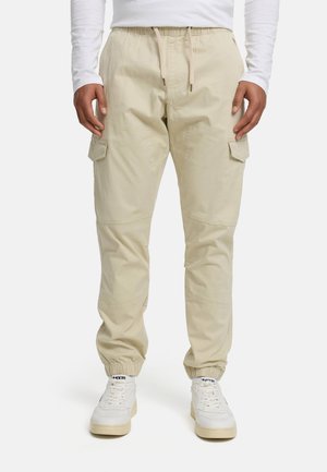 Hombre vestido con pantalones cargo beige con puños elásticos, zapatillas blancas y una camisa blanca de manga larga, de pie mirando hacia adelante.