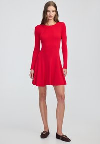 Rode geribbelde gebreide jurk met lange mouwen, aansluitend lijfje, uitlopende rok en ronde halslijn. Gecombineerd met donkerbruine platte schoenen en minimale accessoires.