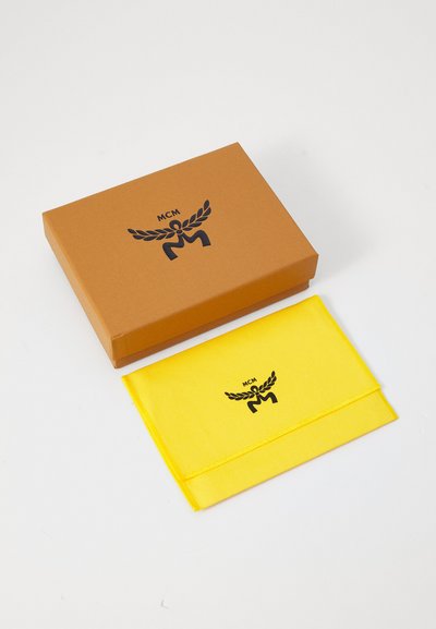 En brun boks med svart MCM-logo ligger ved siden av en gul pose med samme logo, begge på en hvit bakgrunn.