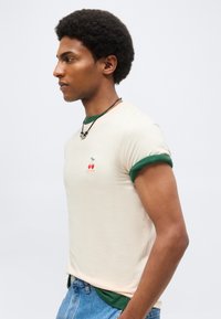 Νέος άνδρας με κρεμ μπλουζάκι με πράσινη λεπτομέρεια και γραφικό κεράσι, φοράει μαύρο κορδόνι κολιέ, μπλε τζιν, κοιτάει αριστερά σε ουδέτερο φόντο.