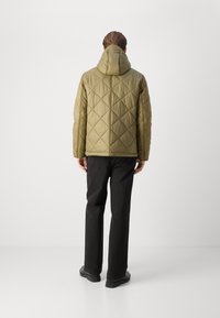 Olivgrüne gesteppte Jacke mit Kapuze, versehen mit einem Rautenmuster. Kombiniert mit schwarzen Hosen und flachen schwarzen Schuhen.