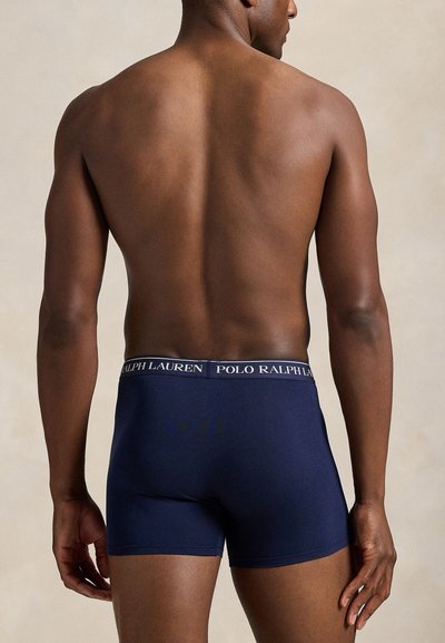 Vyras stovintis nugara į kamerą, dėvintis tamsiai mėlynas Polo Ralph Lauren bokserines trumpikes su firminiu juosmeniu.