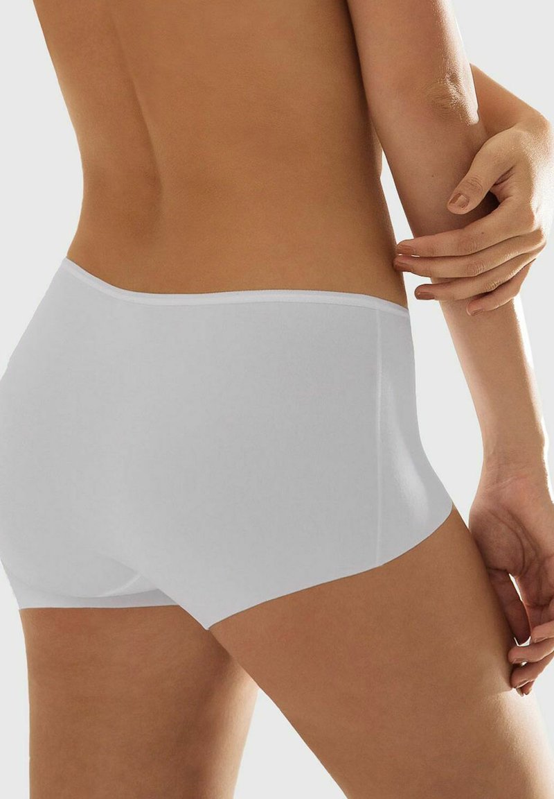 Frau trägt nahtlose, weiße Boyshorts-Unterwäsche, die ihren unteren Rücken und die Oberschenkel zeigt, vor einem neutralen Hintergrund.