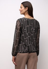 Przezroczysta czarna bluzka z geometrycznym szarym wzorem, uzupełniona elastycznymi rękawami i luźnym krojem. Połączona z beżowymi spodniami.