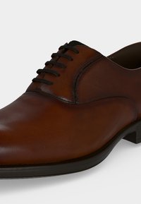 Chaussure habillée en cuir marron avec des lacets noirs et du cuir texturé autour de la zone de laçage, montrée sous un angle trois-quarts.