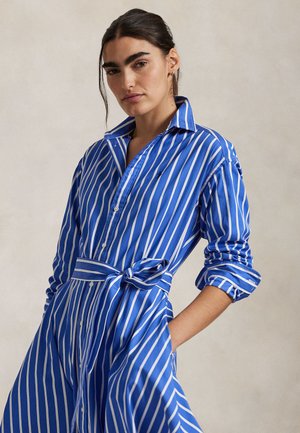 Γυναίκα με σκούρα μαλλιά, φορώντας φόρεμα με κάθετες μπλε και λευκές ρίγες και ζώνη δεμένη στη μέση, στέκεται με το ένα χέρι στην τσέπη μπροστά σε ουδέτερο φόντο.