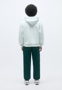 Hoodie grigio chiaro con coulisse e polsini a costine, abbinato a pantaloni verde scuro. Le calzature presentano dettagli in bianco e nero.