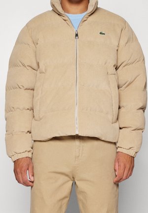 Mężczyzna ubrany w beżową kurtkę puchową z sztruksu z przednim zamkiem błyskawicznym, elastycznymi mankietami i logo Lacoste na piersi, w połączeniu z beżowymi spodniami.