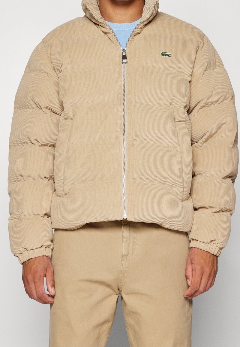 Homme portant une doudoune en velours côtelé beige avec fermeture éclair frontale, poignets élastiques, et logo Lacoste sur la poitrine, associée à un pantalon beige.