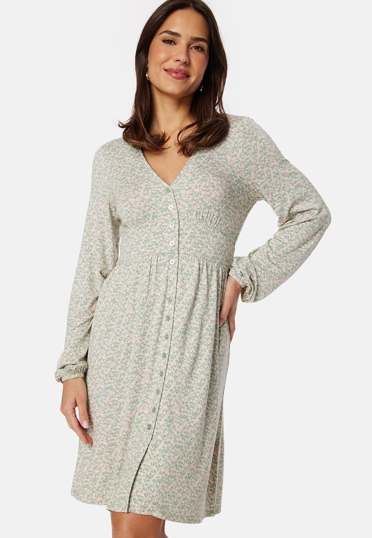Vestito floreale verde chiaro con maniche lunghe, scollo a V, chiusura con bottoni sul fronte e dettagli arricciati in vita. Tessuto morbido con una vestibilità rilassata.