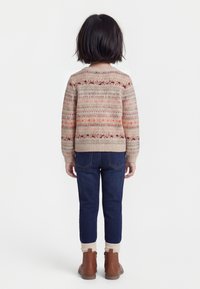 Laps seisab seljaga vaataja poole, seljas mustriline beež kampsun, tumesinised teksad, kreemikad sokid ja pruunid pahkluu saapad valgel taustal.