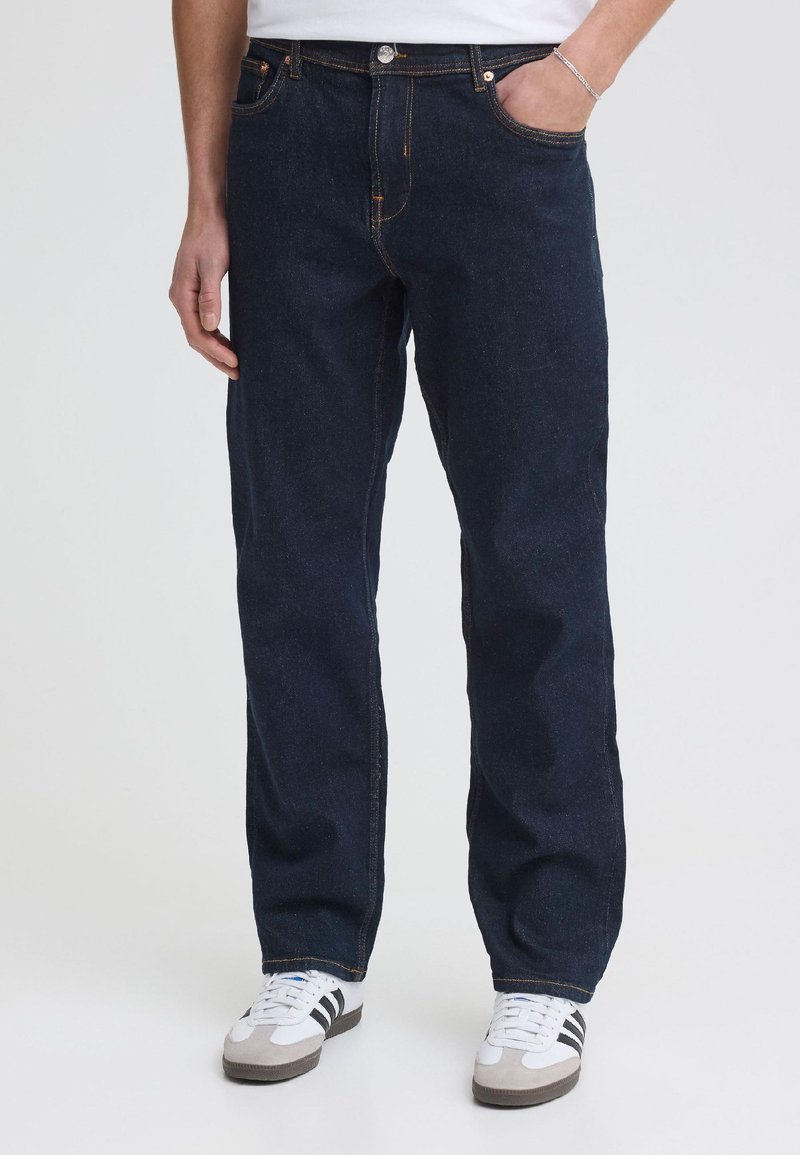 Homme portant un jean droit bleu foncé, une main dans la poche et des baskets blanches avec des rayures noires.