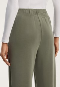 Zelené, lehké a pružné kalhoty s hladkým povrchem a elastickým pasem, zobrazené zezadu, s přiléhavým bílým topem s dlouhým rukávem.