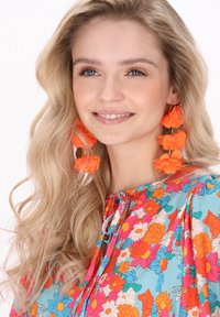 Barevná květinová blouse v tyrkysové barvě, zdobená jasně oranžovými květy a zavazováním u výstřihu. Velké oranžové květinové náušnice doplňují outfit.