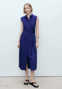 Vestito senza maniche di un blu regale, con chiusura a bottoni frontale, vita annodata, tessuto fluido e spacco anteriore. Abbinato con sandali neri.