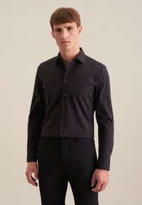 Giovane uomo con capelli corti castani, che indossa una camicia nera aderente e pantaloni neri su uno sfondo beige semplice.