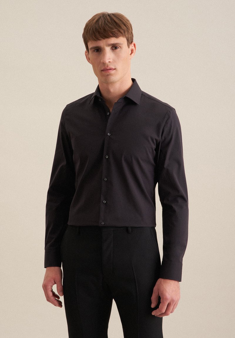 Giovane uomo con capelli corti castani, che indossa una camicia nera aderente e pantaloni neri su uno sfondo beige semplice.