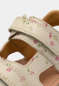 Δερμάτινο σανδάλι ανοιχτού μπεζ χρώματος με floral μοτίβο σε ροζ και πράσινο, διαθέτει δύο λουράκια με βέλκρο και σχέδιο με κοψίματα για καλύτερο αερισμό.