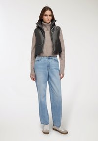 Gilet imbottito nero con colletto alto sopra un dolcevita beige, abbinato a jeans a gamba larga azzurri chiari e sneakers bianche con dettagli color tan.