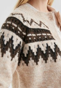 Strickcardigan mit einem strukturierten cremefarbenen Körper und geometrischen Mustern in Dunkelbraun und Schwarz an den Ärmeln und Schultern.