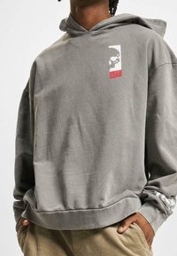 Szara bluza z kapturem, przednią kieszenią, kapturem ze sznurkiem, białą grafiką czaszki i czerwonym akcentem. Charakteryzuje się luźnym krojem i miękką teksturą.