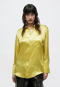 Femeie cu păr lung și întunecat, purtând o cămașă de mătase galbenă lucioasă cu nasturi și pantaloni din piele neagră, stând în fața unui fundal alb simplu.