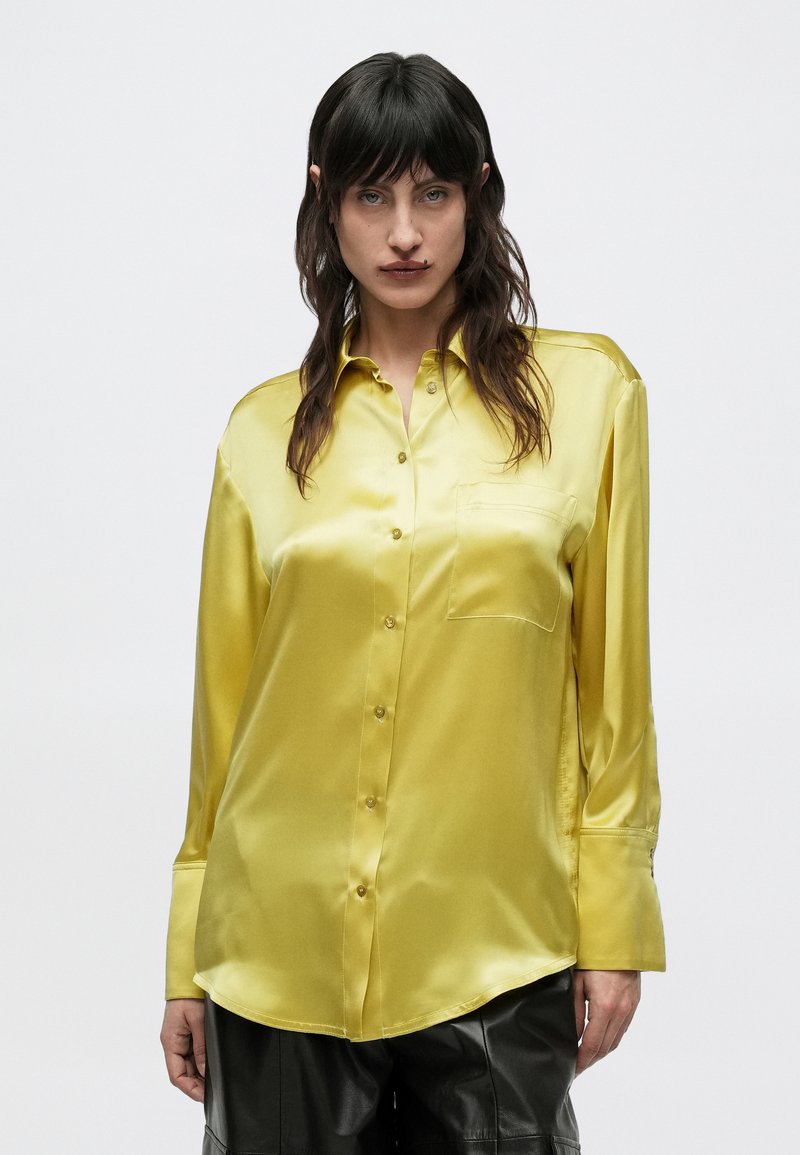 Femeie cu păr lung și întunecat, purtând o cămașă de mătase galbenă lucioasă cu nasturi și pantaloni din piele neagră, stând în fața unui fundal alb simplu.