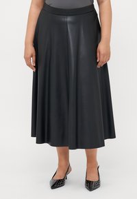 Nainen, jolla on yllään musta tekonahkainen midi-hame, jossa on istuva vyötärö, ja mustat niitillä varustetut kärjelliset slingback-kengät, seisoo valkoista taustaa vasten.
