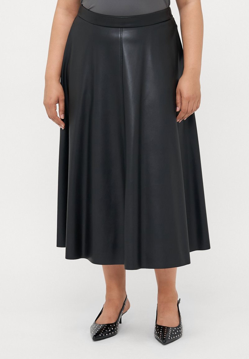 Nainen, jolla on yllään musta tekonahkainen midi-hame, jossa on istuva vyötärö, ja mustat niitillä varustetut kärjelliset slingback-kengät, seisoo valkoista taustaa vasten.