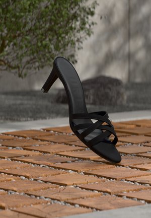 Sandale noire à talons à brides lévitant au-dessus d'une terrasse en bois extérieure avec une plante feuillue et un mur en béton en arrière-plan.
