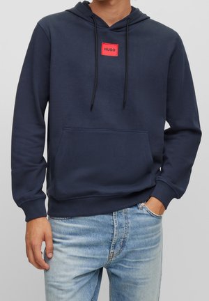 Mężczyzna w granatowej bluzie z kapturem z przednią kieszenią i czerwoną metką "HUGO", w jasnoniebieskich dżinsach, z ręką w kieszeni.