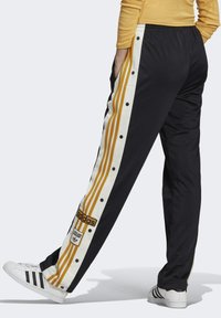 Person som bär svarta Adidas träningsbyxor med gula och vita sidsränder och vita Adidas sneakers, stående med ett ben framåt.