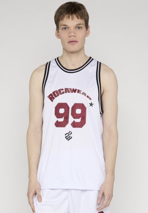 Młody mężczyzna w białej bezrękawnikowej koszulce do koszykówki z napisem "ROCAWEAR 99" w czerwonym i czarnym wykończeniu, stoi na jednolitym tle.