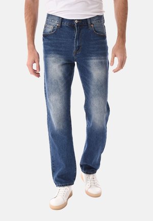 Uomo che indossa jeans blu sbiaditi e sneakers bianche, cammina con le mani rilassate ai lati, su uno sfondo chiaro uniforme.