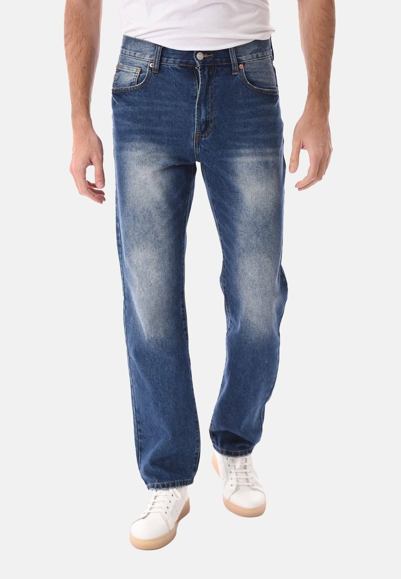 Uomo che indossa jeans blu sbiaditi e sneakers bianche, cammina con le mani rilassate ai lati, su uno sfondo chiaro uniforme.