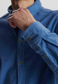 Camisa de botón de color azul claro con mangas largas, que presenta botones de latón, una textura suave y un cuello puntiagudo. El puño muestra un detalle doblado.