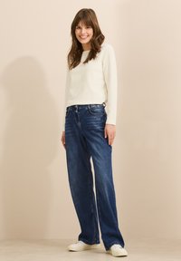 Vit ribbad långärmad topp i kombination med vida blå denimjeans. Modellen bär vita sneakers. Slät textur, avslappnad passform, minimalistisk stil.