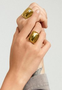 Anillos gruesos de tono dorado en dos dedos, con una superficie suave y reflectante. Las manos tienen tonos de piel claros y un tatuaje visible cerca de la muñeca.