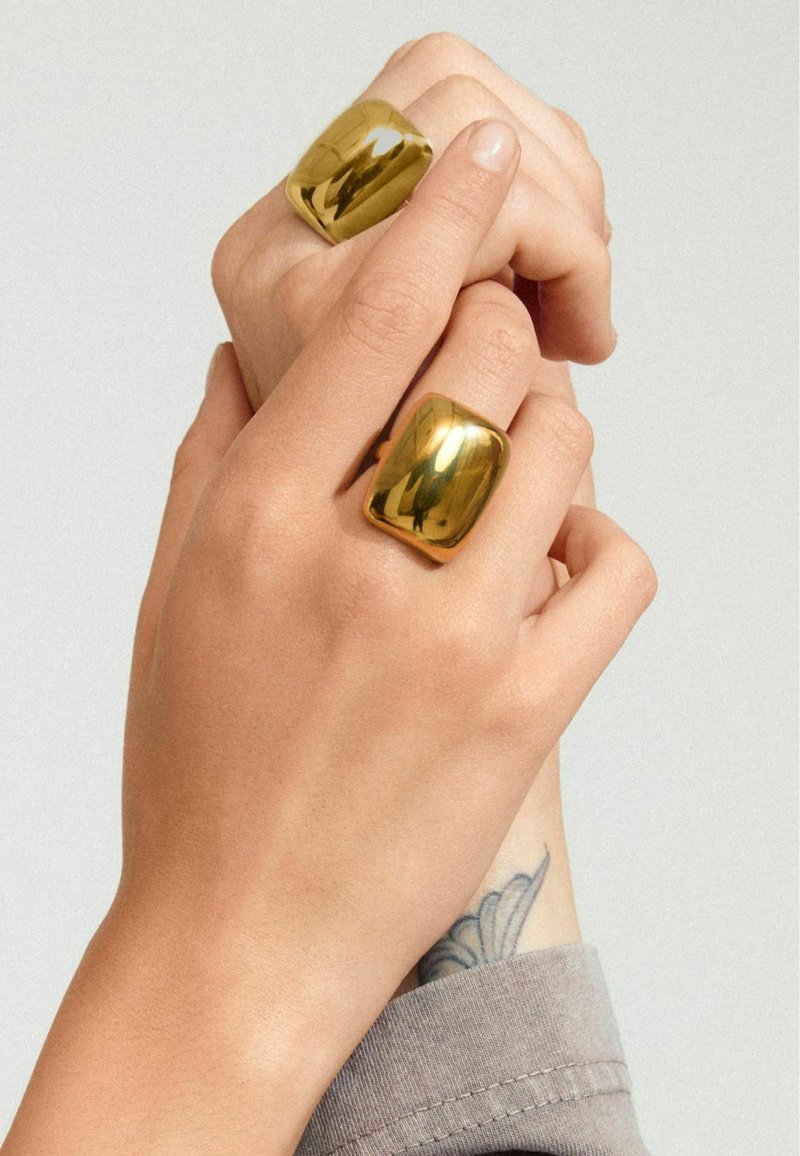 Anillos gruesos de tono dorado en dos dedos, con una superficie suave y reflectante. Las manos tienen tonos de piel claros y un tatuaje visible cerca de la muñeca.