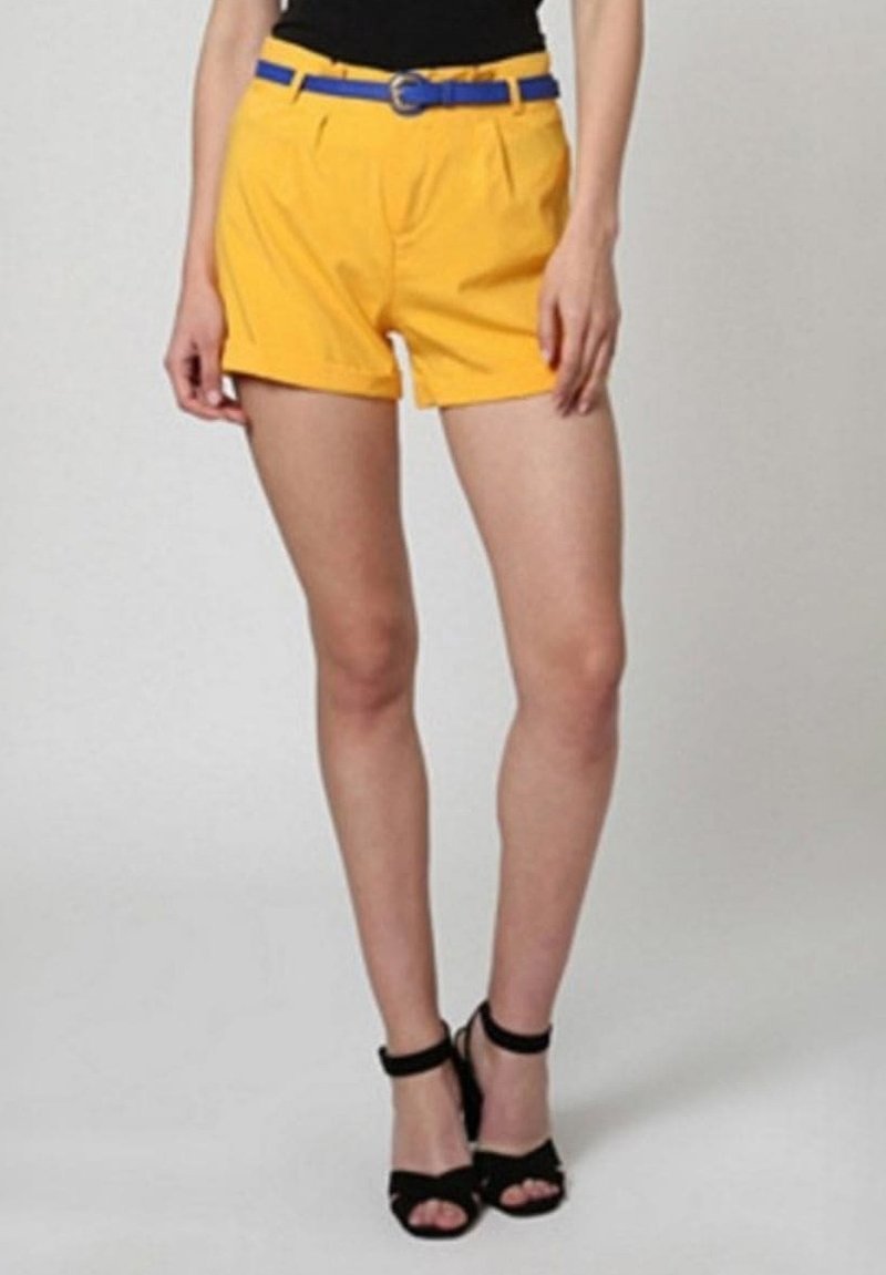 Pantalones cortos amarillos con un cinturón azul, con pliegues en la parte delantera. Combinados con sandalias de tacón alto negras. Fondo liso.