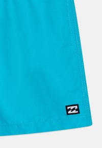 Shorts de bain turquoise en tissu léger, avec des coutures blanches et une étiquette noire avec un logo de vague à l'ourlet.