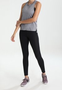 Persona de pie vistiendo una camiseta sin mangas gris de entrenamiento, leggings negros con paneles de malla y zapatillas moradas de Nike sobre un fondo blanco.