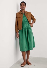 Brun beige jeansjacka som slutar vid midjan över en grön, lager-på-lager, utan ärm-klänning med smockad midja. Bruna sandaler med remmar fulländar outfiten.