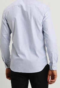 Ljusblå och vit randig långärmad skjorta med rundad nederkant, knäppta manschetter och en mjuk bomullsliknande textur, sedd bakifrån.