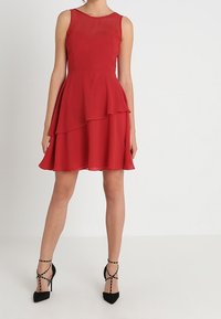 Robe sans manches rouge avec jupe à volants, fabriquée en tissu léger. Texture lisse, longueur genoux, assortie à des talons hauts noirs à brides.