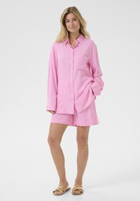 Pink button-up shirt with long sleeves and front pocket, paired with matching pink shorts. Model wearing tan slide sandals.