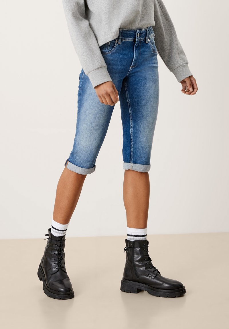 Denim bermudské šortky ve světle až tmavě modrém gradientu s ohrnutím, doplněné černými šněrovacími botami a bílými pruhovanými ponožkami.