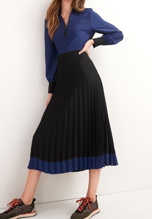 Femme portant un haut bleu à manches longues, une jupe midi plissée noire et des baskets à imprimé léopard avec des lacets orange, debout contre un mur blanc.
