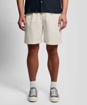 Jambes masculines portant un short beige, des chaussettes blanches et des baskets en toile bleues, debout sur un fond clair uni.